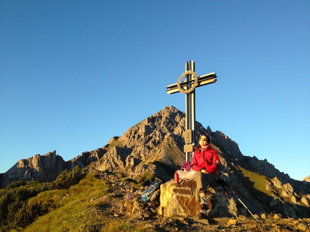 Sonne, Gipfelkreuz und ein Sitzplatz. Im Hintergrund: der Gipfel der Nockspitze.