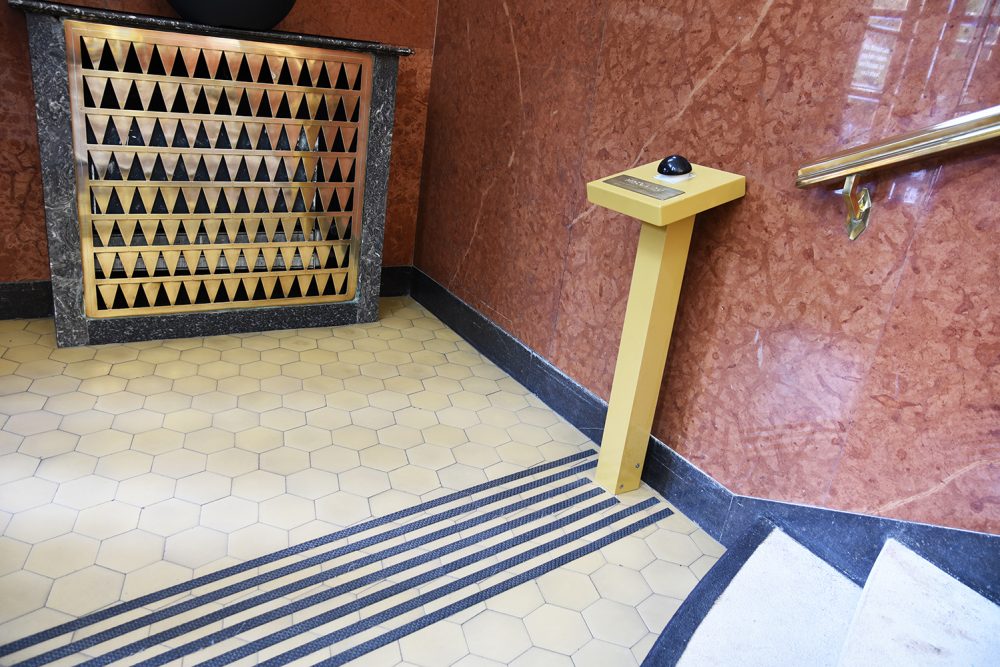 Nella foto a sinistra, dietro la griglia d'ottone, c'è un termosifone. Il tasto nero fa parte del percorso di guida tattile per non vedenti. Foto: Vil Joda.