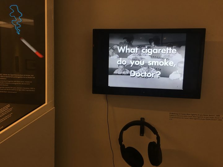 A destra pubblicità della Camel in cui un medico fa da testimonial alla marca di sigarette. Riflessa nel vetro a sinistra l'opera 'Smoke' di Cosima von Bonin. In mostra al Volkskunstmuseum di Innsbruck, Foto © Laura Manfredi